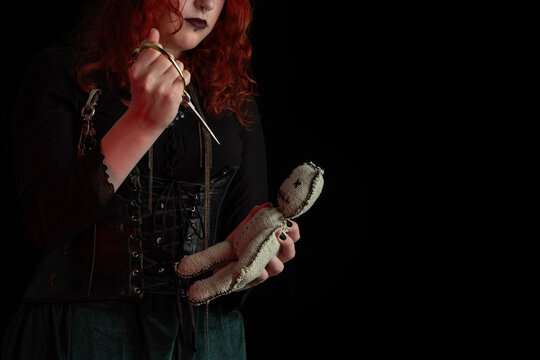 Witch wizard witchcrafting, performing ritual with handmade voodoo doll. Young woman, a magician girl with red hair, dressed in Halloween attire. Halloween night