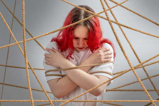 Young female with lowered head trapped in crisscross ropes on gray background, concept of boundaries, rules, stigma, manipulation, isolation, depression and trauma.