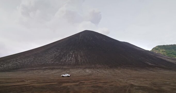 Pick-up truck crossing a dark volcanic ash plain at the base of Mount Yasur on Tanna Island, Vanuatu, under cloudy sky, remote off-road adventure near an active volcano. Tracking side view shot