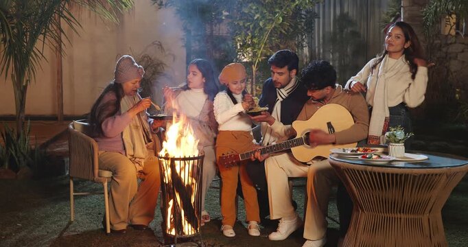 Indian young adult male man sitting on chair hold guitar play music sing song enjoy cold night trip spend time happy joint family female mom group eat tasty food girl child talk speak garden place