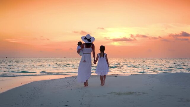 A elegant mother in a white summer dress walks with her children on a tropical paradise beach in the Indian Ocean during sunset time