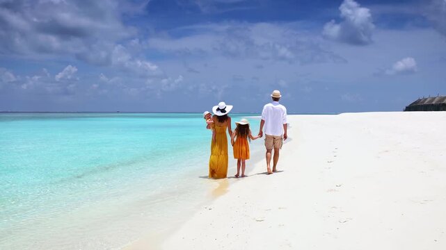 A family of four walks together on a tropical paradise beach with turquoise ocean and white sand during their vacation time in the Maldives islands