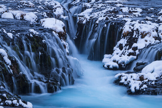 View of the turquoise waters of Bruarfoss waterfall cascading through dark volcanic rocks covered in a thick layer of fresh white snow in Iceland.