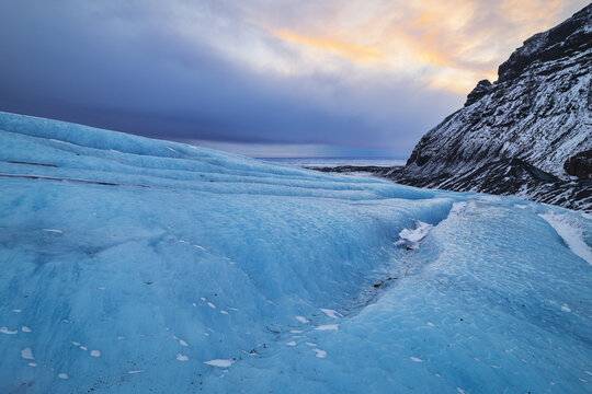 View of the blue ice of Falljokull glacier with deep crevasses under a sunset sky next to a snow-dusted mountain ridge Sveitarfelagid Hornafjordur, Iceland.
