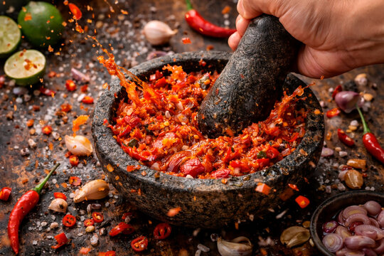 Dynamic splash of fresh sambal being crushed in a traditional stone mortar and pestle (cobek) with red chili peppers, garlic, and lime.