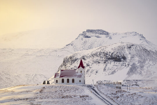 View of the historic white church with a red roof nestled against snow-covered mountains in Eyja- og Miklaholtshr, Iceland.