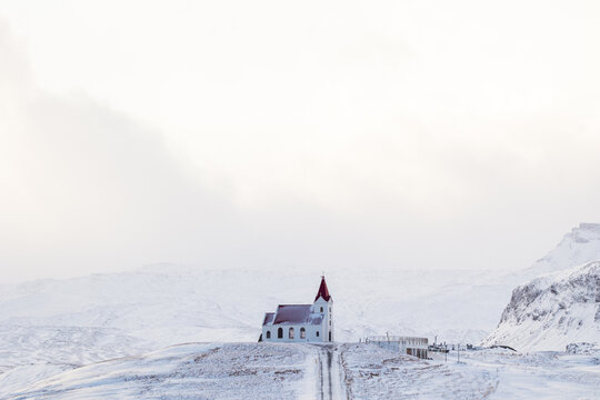 View of a small white church with a red roof standing on a snowy hill against a backdrop of rugged mountains in Eyja- og Miklaholtshr, Iceland.