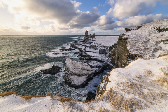 View of the Snaefellsnes coastline featuring snow-covered volcanic sea stacks and crashing waves under a cloudy sky Eyja- og Miklaholtshr, Iceland.