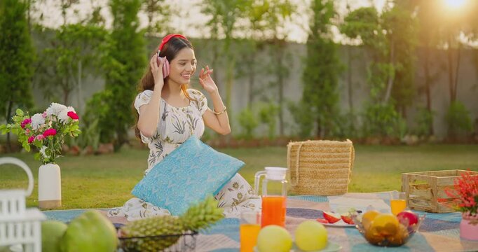 Indian gen z female sitting on mat wear headset listen good mp3 music sing song enjoy day fresh air garden Happy young adult relax girl dancing spend time feel free have fun joy at picnic trip place