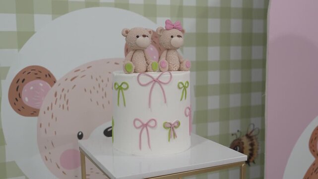A round cake sits on a table with two teddy bear figures on top. The cake has pink and green decorations. The background shows cheerful patterns and colors for a birthday celebration.