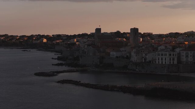 Aerial view of Antibes, France, as a slow sweep reveals old town, Chateau Grimaldi, medieval ramparts, beaches, boats, people, and calm Mediterranean at sunset.
