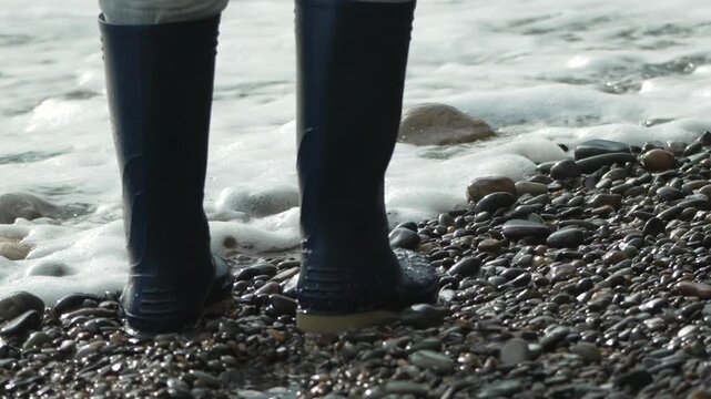 Boots, pebble, beach, person walks in rubber waders on wet shingle shore, then kneels down near washing sea foam.