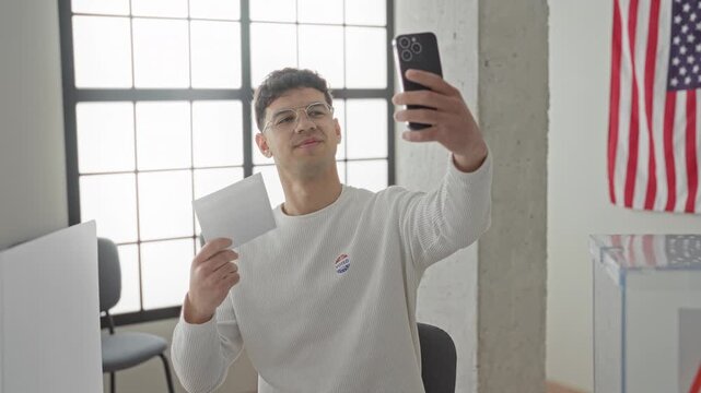 Young hispanic man holding ballot envelope and smartphone, raising phone for selfie at a polling station; civic pride.