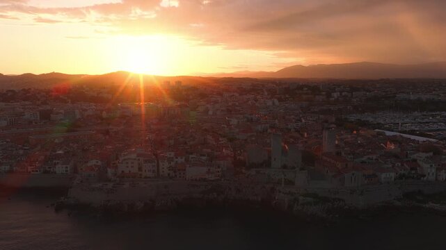 Aerial view over Antibes, France, showing rocky headland, medieval ramparts, Chateau Grimaldi, terracotta roofs, and a busy marina at sunset.