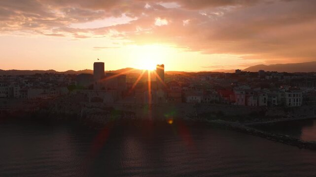 Aerial view of Antibes, France, with castle towers, flag, rocky ramparts and a marina, as the sun sets, lens flares sparkle, reflections ripple on calm sea.