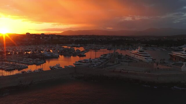 Aerial view over Antibes shows Port Vauban and old ramparts at sunset. A slow glide reveals yachts, masts, and orange reflections, with hills in silhouette.