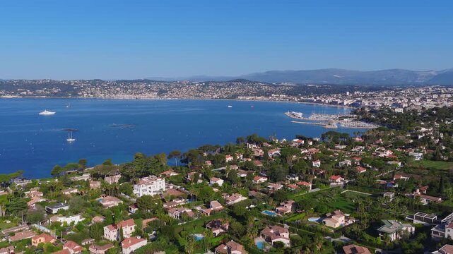 Aerial view over Antibes on the French Riviera shows curving bay, Cap d'Antibes, yacht marina, breakwater, villas, pine trees, pools, old town, and Chateau Grimaldi.