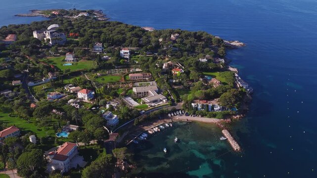 Aerial high angle drone sweep shows Cap d'Antibes in France, rocky coast, tree lined estates, castle like villa, moored yachts, stone breakwater, pools, and turquoise shallows.