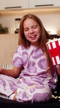 Teenager girl at night home holds popcorn bowl watches movie laughs drops snack on seat starts clean. Caucasian child kid on sofa at evening picks pieces by hand wipes crumbs smiles enjoys fun tidy