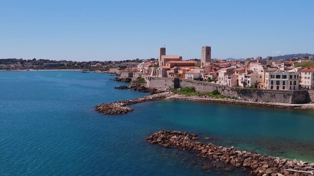 Aerial view glides along Antibes France, showing walled old town, medieval towers, terracotta roofs, rocky ramparts, coves and breakwaters under a clear midday sky.