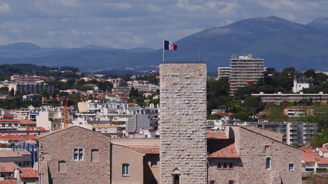 Aerial panorama of Antibes, France shows a medieval stone tower with a French flag, terracotta roofs, mid century blocks, wooded hills, and distant mountains as the camera sweeps