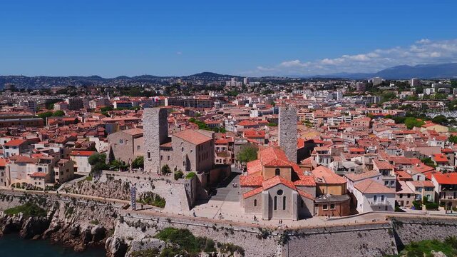 Aerial view of Antibes on the French Riviera shows old walled town, twin towers, red tiled church, terracotta roofs, cobbled streets, promenade, marinas, yachts.