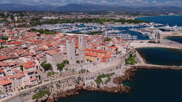 Aerial view over Antibes, France shows Vieil Antibes walls, twin keeps, Chateau Grimaldi, Port Vauban, sandy beach, red tiled roofs, boats, bathers, pedestrians.