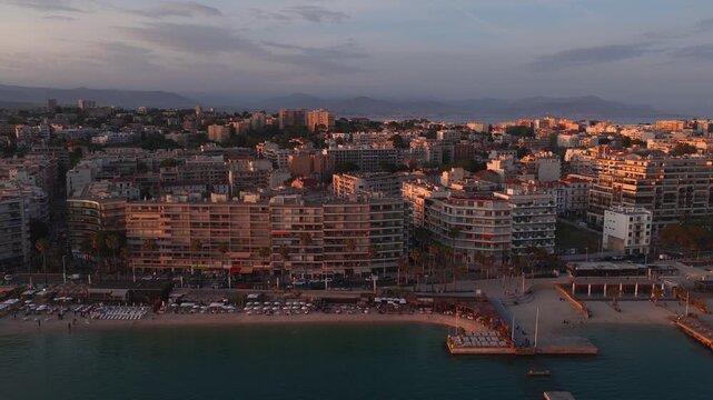 Aerial view of Antibes, France at sunset shows sandy beach, rows of umbrellas, concrete piers, a small boat by the jetty, palm promenades and urban blocks in warm light.