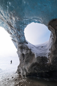 View of a person standing on a snowy plain seen through a circular opening in the blue ice of the Svinafellsjokull glacier cave, Sveitarfelagid Hornafjordur, Iceland.