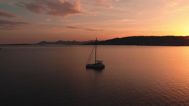 Aerial sunset over Antibes on the French Riviera shows a catamaran at anchor, a starburst sun behind the headland, a golden reflection, yachts, beaches, and calm ripples.