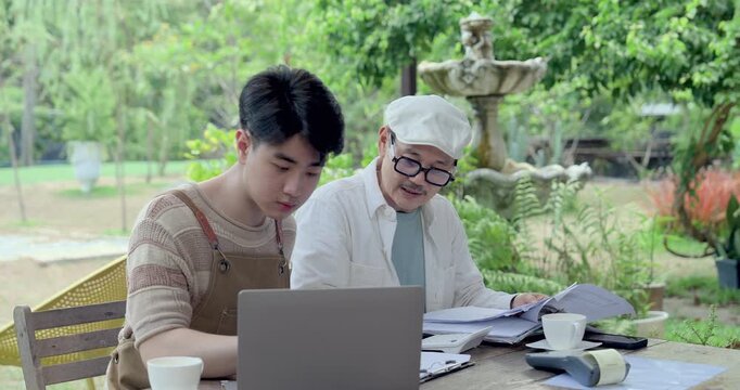 Asian father and son review laptop screen, paperwork, invoice folder, coffee cup, planning cafe daily task, order update, accounting summary, family business mentorship and guidance session.