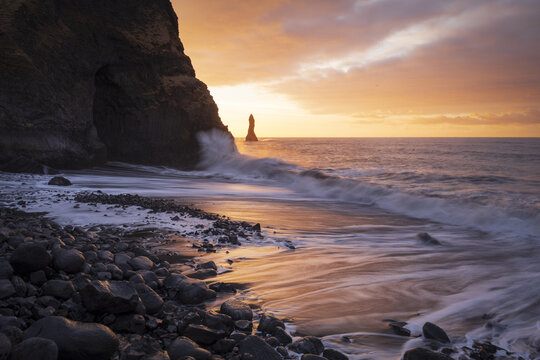 View of the Reynisdrangar sea stacks and Reynisfjara black sand beach with waves crashing against basalt cliffs under a golden sunset in Vik, Myrdalshreppur, Iceland.