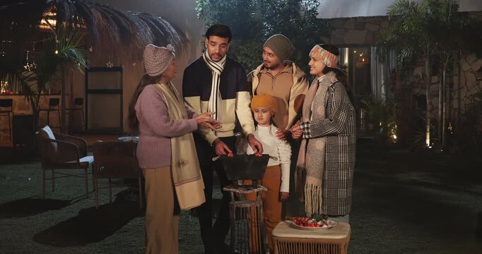 Indian happy mature adult female male speak share gossip enjoy winter cold have fun joy night outdoor garden place mom girl child kid talk with young man cook party food on barbecue spend quality time