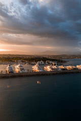 Fototapeta premium Aerial view of Port Vauban in Antibes, France, with rows of superyachts along the breakwater at sunset. The harbor curves to the old town, with Alps faintly visible.
