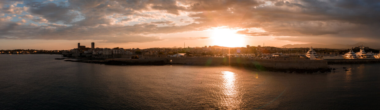Aerial sunset view of Antibes, France, with Bay of Angels, old town seawall, Chateau Grimaldi towers, and yachts at Port Vauban, warm clouds, calm Mediterranean waters.