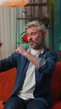 Trendy cheerful positive middle-aged man having fun dancing and moving to rhythm, dabbing raising hands, making dub dance gesture at modern home apartment. Mature senior guy in living room on sofa