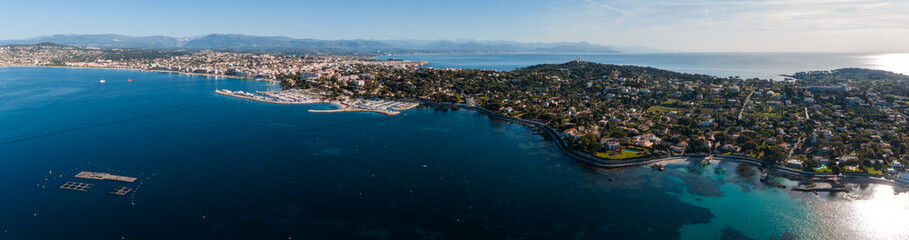 Fototapeta premium Aerial view of Antibes, France, with Cap d Antibes curving into the sea, old town and harbor on the left, Fort Carre and marina basins, Alps on the hazy horizon.