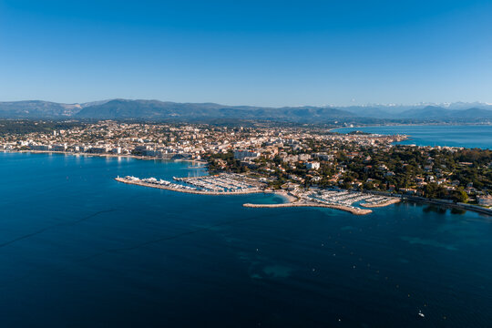 Aerial scene of Antibes, France, shows Cap d Antibes, marinas with yachts, sandy beaches, curving harbor basins, old town and Alps in late afternoon golden light.