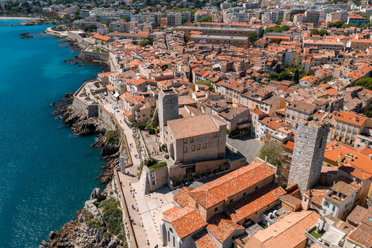 Aerial view of Antibes, France, shows old town stone walls, red tiles, and the square bell tower of Chateau Grimaldi by the seafront. Golden hour light, people stroll the path.