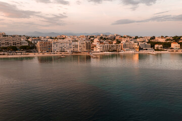 Fototapeta premium Aerial view of Antibes, France, at sunset shows modern mid rise hotels and apartments by a calm bay, old town cluster, sandy beaches, pastel clouds, and distant hills.