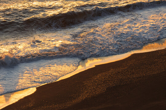 View of waves crashing on the Reynisfjara black sand beach with golden sunlight reflecting on the water and foam in Vik, Myrdalshreppur, Iceland.