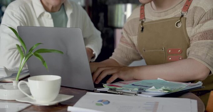 Close view of son using laptop beside cafe paperwork, father nearby supporting family restaurant inventory update, sales report review, accounting document, operation planning task with chart