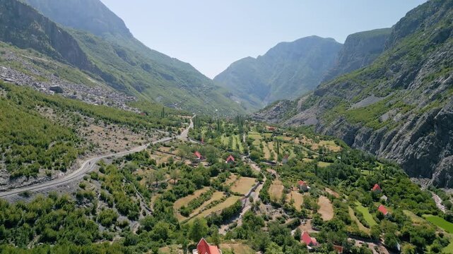 Aerial forward and rising drone flight over the scenic SH20 mountain road descending into a green valley with houses in the Albanian Alps. Clear blue sky and rocky mountain peaks