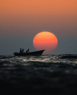 View of fishermen in a small boat silhouetted against a massive orange sun setting over the calm ocean waves Balochistan, Pakistan.
