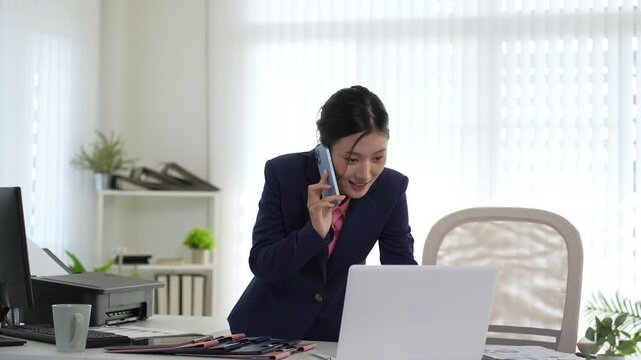 Asian businesswoman professional talking phone call working laptop computer standing office desk multitask communication corporate workplace modern bright