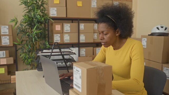 Woman holding a parcel and red pen while sorting stacked packages and working on a laptop at a packing desk in building; small business focus.