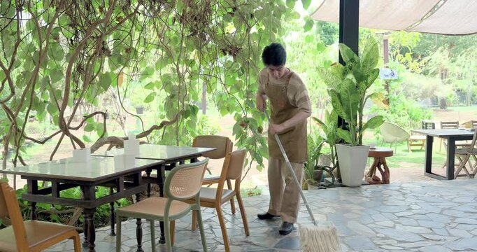 Asian son mops garden cafe floor between table and chair, practicing cleanliness standard, hygiene routine, maintenance duty, family restaurant operation training, responsible service preparation.