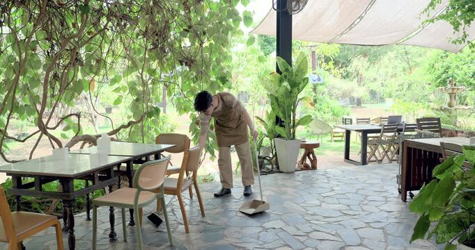 Asian son sweeps patio floor beside cafe table and chair, practicing hygiene routine, housekeeping duty, service readiness, family restaurant training, maintenance skill for daily operation.