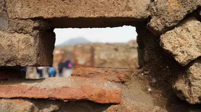 View through a small Pompeii wall opening with rough stone and brick in focus, framing blurred unrecognizable visitors and distant ruins beyond.
