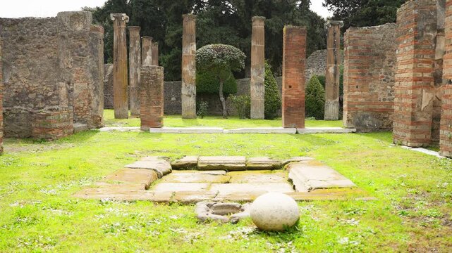 Wide view of a Pompeii peristyle courtyard with standing columns, brick supports, lawn, and a central stone feature, showing the open layout of an ancient Roman house.